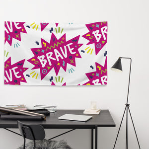 A large flag is featured hanging on the wall of an office placed above a desk. The white flag features a pattern with the word "brave" featured inside a pink star-like shape with sketchy illustrations around the word. 