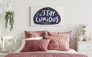 A large flag featured on a wall of a girls bedroom above the bed. The white flag features the words "stay curious" featured inside a a purple shape with sketchy illustrations around. 