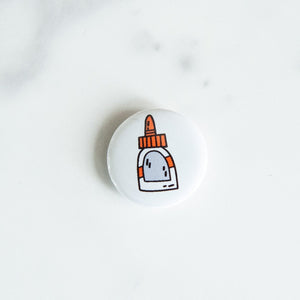 A button pin on a white tabletop. The button features an illustration of a glue bottle. 