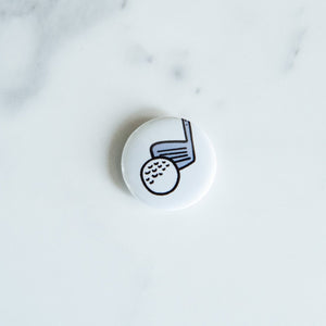 A buttons on a white tabletop. The button features an illustrations of a golf club with ball. 