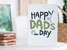 Load image into Gallery viewer, A greeting card featured standing up on a white tabletop with a framed photo of a succulent in the background and a stack of wooden coasters. There’s a woven basket in the background with a cactus inside. The card features the words “Happy Dad’s Day” with an illustrated golf club and golf ball. 