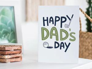 A greeting card featured standing up on a white tabletop with a framed photo of a succulent in the background and a stack of wooden coasters. There’s a woven basket in the background with a cactus inside. The card features the words “Happy Dad’s Day” with an illustrated golf club and golf ball. 