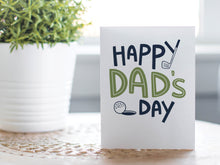 Load image into Gallery viewer, A photo of a card featured on a tabletop next to a white planter filled with a green plant. ​​The card features the words “Happy Dad’s Day” with an illustrated golf club and golf ball. 