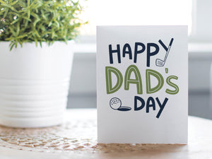A photo of a card featured on a tabletop next to a white planter filled with a green plant. ​​The card features the words “Happy Dad’s Day” with an illustrated golf club and golf ball. 