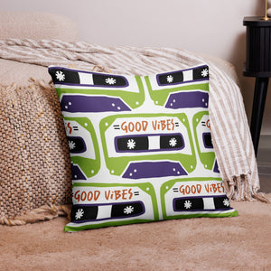 A pillow on the floor on a rug laying against a bed. The white pillow features the artwork on a white background with an illustrated cassette tape pattern with the words 'good vibes.'