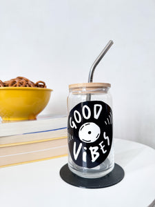 A photo of a glass can tumbler on a coffee table with books and a bowl of pretzels in the background. The glass tumbler has a bamboo lid and silver metal straw. The design on the glass tumbler says the words "good vibes" inside an illustrated vinyl record.