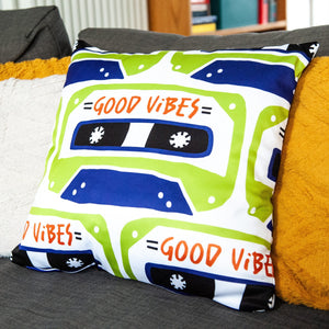 A white pillow on a grey sofa with other pillows. The white pillow features the artwork on a white background with an illustrated cassette tape pattern with the words 'good vibes.'