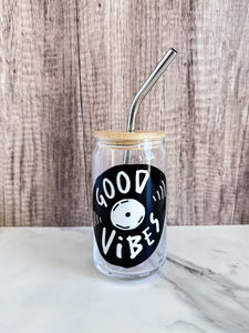 A glass can tumbler featured on a marble tabletop with a wood background. The design on the glass tumbler says the words "good vibes" inside an illustrated vinyl record.