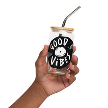 Load image into Gallery viewer, A photo of a glass can tumbler being held in a hand. The glass tumbler has a bamboo lid and silver metal straw. The design on the glass tumbler says the words &quot;good vibes&quot; inside an illustrated vinyl record.