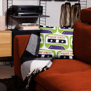 A white pillow on a red sofa. The white pillow features the artwork on a white background with an illustrated cassette tape pattern with the words 'good vibes.'