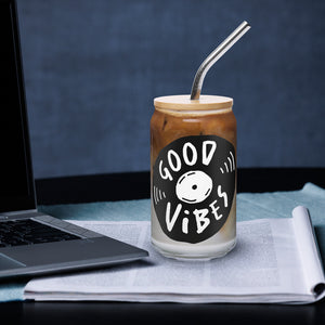 A photo of a glass can tumbler on a desktop next to a laptop. The glass tumbler has a bamboo lid and silver metal straw. The design on the glass tumbler says the words "good vibes" inside an illustrated vinyl record.