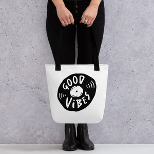 A person holding a white tote bag with black handles. The bag features a design with the words 'good vibes' inside an illustrated vinyl record. 