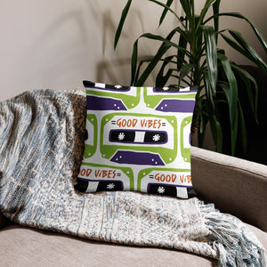 A pillow on a sofa with a blanket. The white pillow features the artwork on a white background with an illustrated cassette tape pattern with the words 'good vibes.'