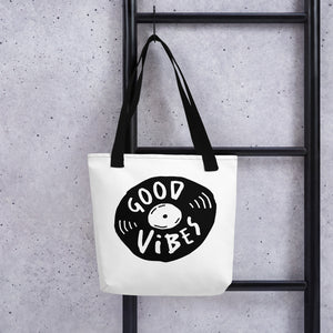 A white tote bag with black handles handing on a black ladder. The bag features a design with the words 'good vibes' inside an illustrated vinyl record. 