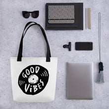 Load image into Gallery viewer, A white tote bag with black handles on a grey tabletop with a laptop and other office supplies. The bag features a design with the words &#39;good vibes&#39; inside an illustrated vinyl record. 