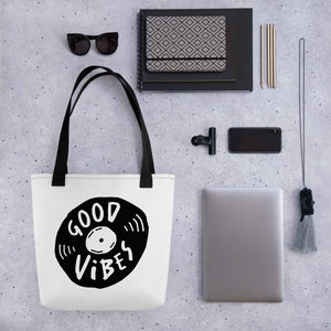 A white tote bag with black handles on a grey tabletop with a laptop and other office supplies. The bag features a design with the words 'good vibes' inside an illustrated vinyl record. 