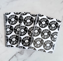 Load image into Gallery viewer, Three spiral notebooks are on a tabletop. The white spiral notebooks have black spiral binding. The pattern design features the words &quot;good vibes&quot; inside an illustrated vinyl record.  