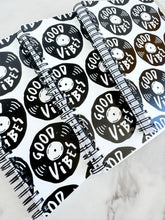 Load image into Gallery viewer, Three spiral notebooks are on a tabletop. The white spiral notebooks have black spiral binding. The pattern design features the words &quot;good vibes&quot; inside an illustrated vinyl record.  