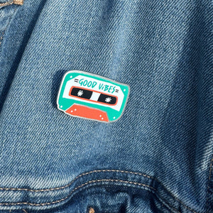 An enamel pin featuring the words "good vibes" inside an illustrated cassette tape. The pin is on a jean jacket.