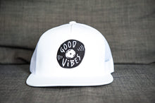 Load image into Gallery viewer, A white trucker hat sitting on a gray sofa. The hat features an black embroidered design with an illustrated vinyl record and the words &#39;good vibes&#39; inside.  