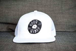 A white trucker hat sitting on a gray sofa. The hat features an black embroidered design with an illustrated vinyl record and the words 'good vibes' inside.  