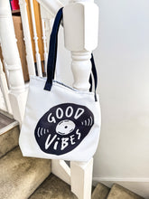 Load image into Gallery viewer, A white tote bag with black handles hanging on a staircase. The bag features a design with the words &#39;good vibes&#39; inside an illustrated vinyl record. 