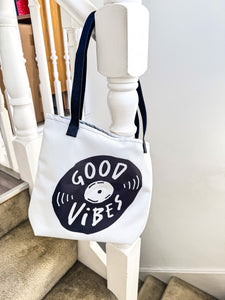 A white tote bag with black handles hanging on a staircase. The bag features a design with the words 'good vibes' inside an illustrated vinyl record. 