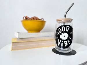 A photo of a glass can tumbler on a coffee table with books and a bowl of pretzels in the background. The glass tumbler has a bamboo lid and silver metal straw. The design on the glass tumbler says the words "good vibes" inside an illustrated vinyl record.