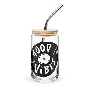 A photo of a glass can tumbler on a white background with a bamboo lid and silver metal straw. The design on the glass tumbler says the words "good vibes" inside an illustrated vinyl record.