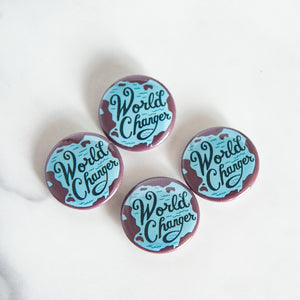 Buttons on a white tabletop. The buttons feature word art illustrations with the words "World Changer."