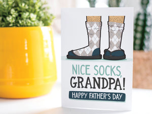 A greeting card is on a table top with a yellow plant pot and a green plant inside. The card features the words “Nice Socks Grandpa, Happy Father’s Day” with an illustrated of legs with patterned socks and shoes. 