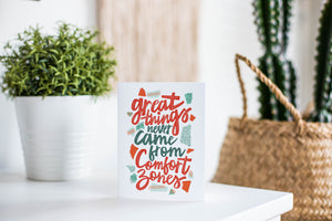 A greeting card featured standing up on a white tabletop with a pink plant pot in the background and some succulents in the pot. There’s a woven basket in the background with a cactus inside. The card features the words “Great things never came from comfort zones.”