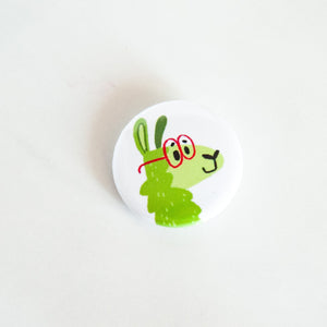 A buttons on a white tabletop. The button features a green illustrated llama with red glasses on. 