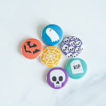 Load image into Gallery viewer, A set of six buttons on a white tabletop. The buttons feature illustrations of a ghost, bats, skeletons, a skull, a grave, and a spiderweb.