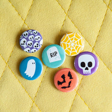 Load image into Gallery viewer, A set of six buttons on a yellow background. The buttons feature illustrations of a ghost, bats, skeletons, a skull, a grave, and a spiderweb.