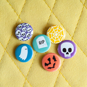 A set of six buttons on a yellow background. The buttons feature illustrations of a ghost, bats, skeletons, a skull, a grave, and a spiderweb.