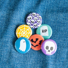 Load image into Gallery viewer, A set of six buttons on a jean jacket. The buttons feature illustrations of a ghost, bats, skeletons, a skull, a grave, and a spiderweb.