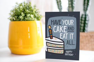 A greeting card is on a table top with a yellow plant pot and a green plant inside. The card features the words “Have Your Cake and Eat it All Happy Birthday” with an illustrated piece of cake with a candle in it.