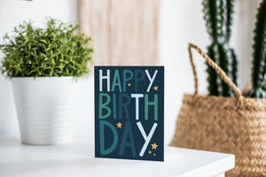 A greeting card is featured on a white tabletop with a white planter in the background with a green plant. There’s a woven basket in the background with a cactus inside. The card features the words “happy birthday” with stars surrounding the letters.