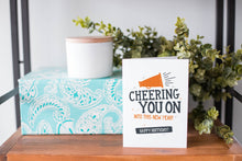 Load image into Gallery viewer, A greeting card is on a table top with a present in blue wrapping paper in the background. On top of the present is a candle and some greenery from a plant too. The card features the words “Cheering You on into This New Year, Happy Birthday.&quot;