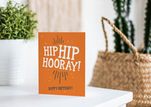 Load image into Gallery viewer, A greeting card is featured on a white tabletop with a white planter in the background with a green plant. There’s a woven basket in the background with a cactus inside. The card features the words “Hip Hip Hooray, Happy Birthday.”