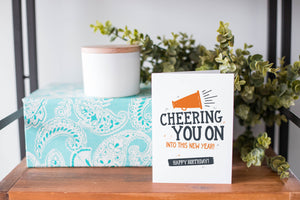 A greeting card is on a table top with a present in blue wrapping paper in the background. On top of the present is a candle and some greenery from a plant too. The card features the words  "Cheering you on into this new year! Happy birthday!."