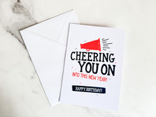 Load image into Gallery viewer, A card with a white envelope laying on a tabletop. The card features the words “Cheering You on into This New Year, Happy Birthday.&quot;