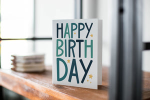 A card on a wood tabletop with an object in the background that is out of focus. The card features the words “Happy birthday” with blue letters featured on a white background. 