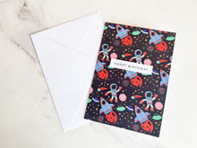 Load image into Gallery viewer, A greeting card laying on a table with a white envelope. The greeting card features the words &#39;happy birthday&#39; with illustrated space items around it. The space items include an astronaut, space rockets, planets and stars. 