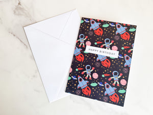 A greeting card laying on a table with a white envelope. The greeting card features the words 'happy birthday' with illustrated space items around it. The space items include an astronaut, space rockets, planets and stars. 