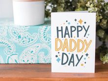 Load image into Gallery viewer, A greeting card is on a table top with a present in blue wrapping paper in the background. On top of the present is a candle and some greenery from a plant too. The card features the words  “Happy Daddy Day” with diamond shapes surrounding the letters. 