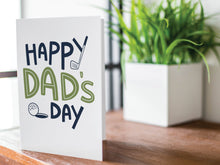 Load image into Gallery viewer, A greeting card is featured on a wood coffee table with a green plant in a white planter in the background. The card features the words “Happy Dad’s Day” with an illustrated golf club and golf ball. 