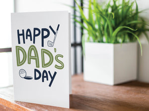 A greeting card is featured on a wood coffee table with a green plant in a white planter in the background. The card features the words “Happy Dad’s Day” with an illustrated golf club and golf ball. 