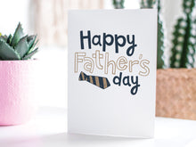 Load image into Gallery viewer, A greeting card featured standing up on a white tabletop with a pink plant pot in the background and some succulents in the pot. There’s a woven basket in the background with a cactus inside. The card features the words “Happy Father’s  Day” with a striped tie on the bottom of the words. 